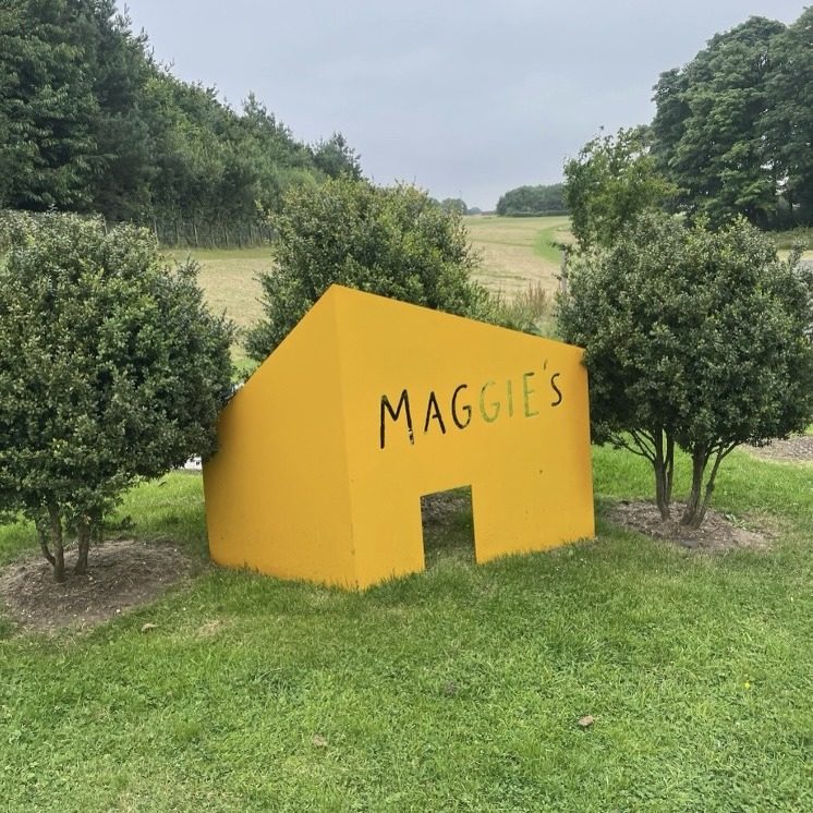 A large yellow house-shaped sign reading Maggie's, standing on grass among shrubs outside the centre.