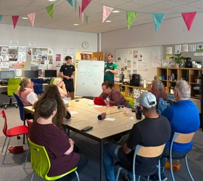 Students leading a discussion with a seated group, with a whiteboard behind them.